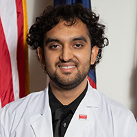 Man with dark, curly hair and short goatee wearing a black shirt, black tie, and white coat with UT Southwestern logo